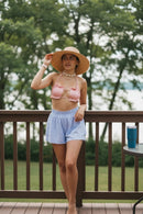 Woman in a pink bikini top and light blue shorts standing on a wooden deck with trees in the background.
