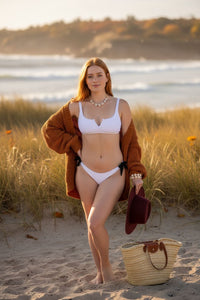 Woman in a white bikini standing on a sandy beach with grass and water in the background